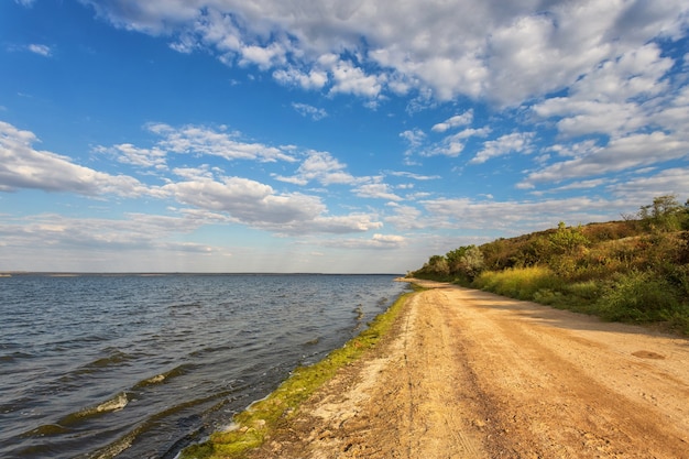 Foto el camino a lo largo de la orilla del lago, río, contra un cielo azul con nubes belvmi