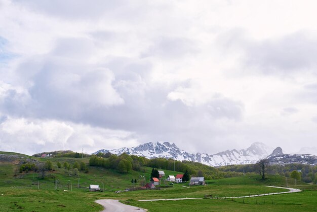 Camino en un pasto verde cerca de una pequeña aldea agrícola al pie de las montañas cubiertas de nieve