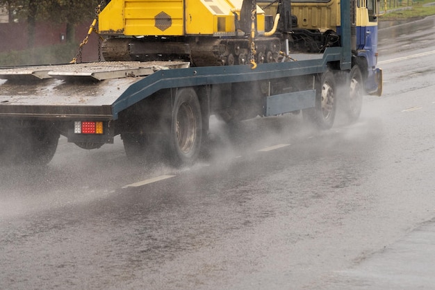 Camión de plataforma corta que lleva un tractor de orugas en una carretera mojada con salpicaduras durante el día lluvioso