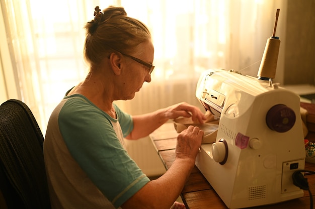 Camisa branca de costureira mulher idosa sênior confiante e óculos, sentado na frente da máquina de costura, trabalhando em roupas em casa usando tecido bege.