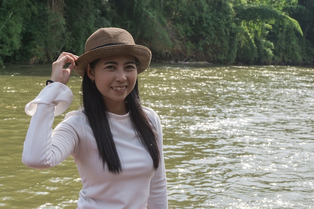 Camisa feliz niña asiática blanca y sombrero marrón en el río en el fondo de la naturaleza