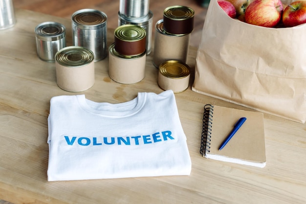 Camiseta branca com inscrição voluntária azul latas saco de papel com maçãs e caderno em madeira