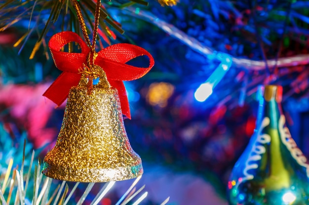 Campana de Navidad dorada con cinta roja contra la guirnalda de colores luminosos en la rama de abeto decorativo de cerca