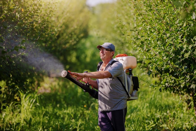Campesino caucásico maduro en ropa de trabajo, sombrero y con la moderna máquina de pulverización de pesticidas en la espalda rociando insectos en el huerto.
