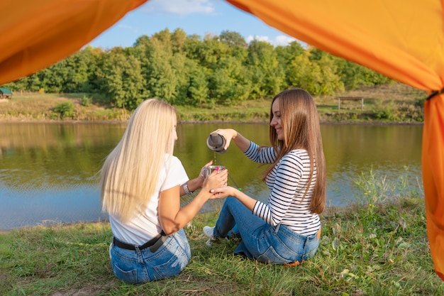 Camping am Seeufer bei Sonnenuntergang, Blick aus dem Zelt. Zwei wunderschöne Mädchen genießen die Natur und trinken heißen Tee vor dem Zelt