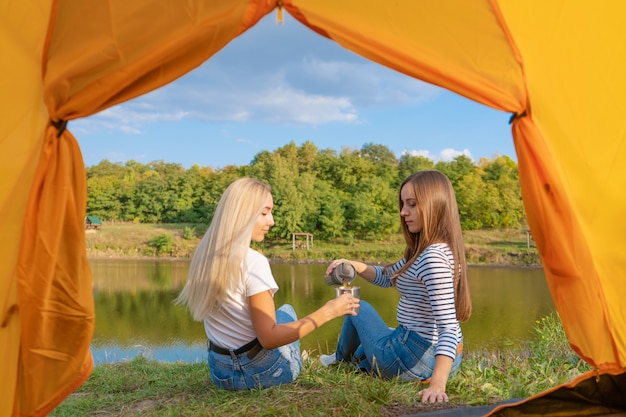 Camping am Seeufer bei Sonnenuntergang, Blick aus dem Zelt. Zwei wunderschöne Mädchen genießen die Natur und trinken heißen Tee vor dem Zelt