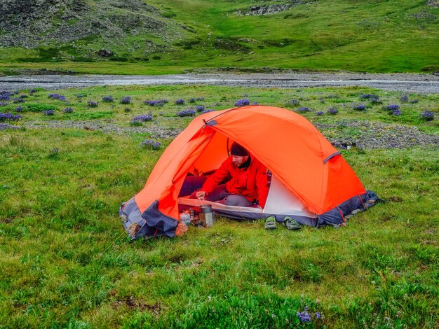 Camping auf einem sommergrünen Hochplateau. Mann in einem orangefarbenen Zelt bereitet Essen zu. Ruhe und Erholung in der Natur. Solo-Wanderkonzept.