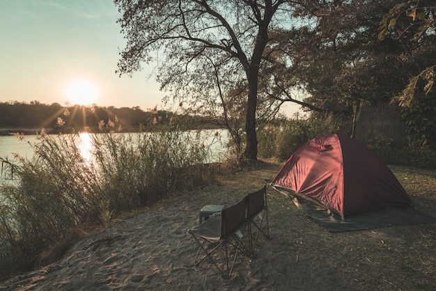 Camping con carpa, sillas y equipo de campamento. salida del sol sobre el río okavango, frontera de namibia botswana. viajes de aventura y actividades al aire libre en áfrica. imagen tonificada, estilo vintage.