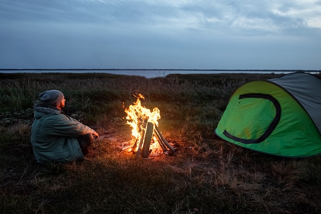 Camping homem sentado perto do fogo à noite contra o céu. viagens, turismo, camping.