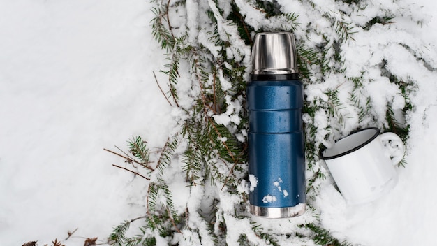 Camping im Winterwaldkonzept. Thermoskanne und Eisenbecher liegen auf einem Kiefernzweig im Schnee. Ansicht von oben, Kopienraum. Isolierflasche für unterwegs.