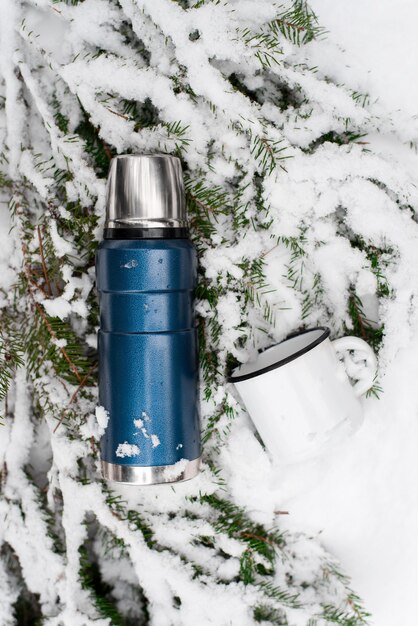 Camping im Winterwaldkonzept. Thermoskanne und lackierter Becher, der auf Kiefernzweig im Schnee liegt. Ansicht von oben, vertikaler Winkel. Isolierflasche für unterwegs