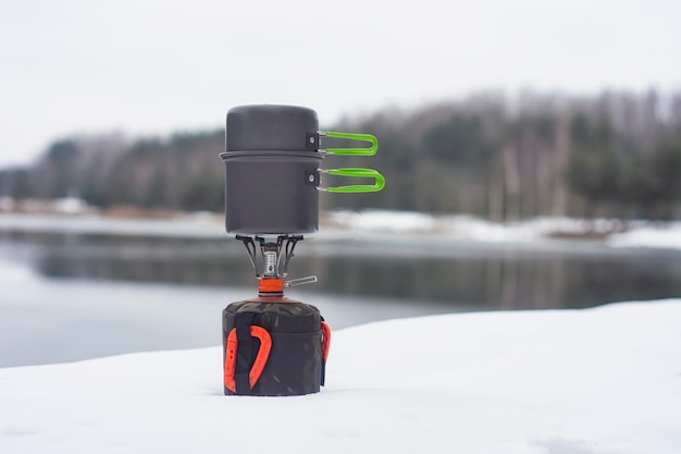 Camping de invierno al aire libre contra el fondo de la nieve del bosque y el lago Quemador de gas turístico con sartenes para cocinar