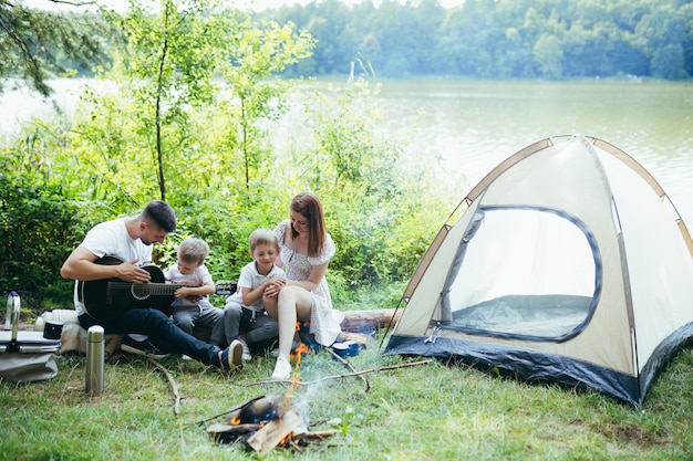 Camping junto al lago en el bosque. familia feliz papá mamá y niños pequeños sentados junto al fuego y la carpa en la naturaleza. pasar tiempo libre juntos de vacaciones. al aire libre. Padres con hijos. padre tocando la guitarra. Acampar