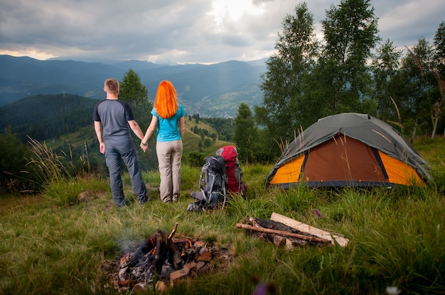 Cámping. Pareja amorosa de pie cerca del fuego, mochilas y carpa