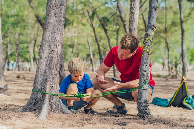 Camping personas estilo de vida al aire libre turistas en el bosque de verano cerca del mar lazur Niño rubio hijo con padre estudiar técnicas de supervivencia practicar métodos de atar nudos de cuerda Educación natural de los niños