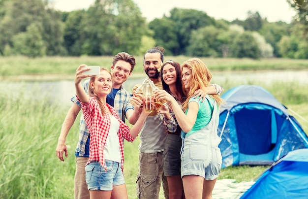 Camping-, Reise-, Tourismus-, Wanderungs- und Personenkonzept - glückliche Freunde mit Glasflaschen, die Apfelwein oder Bier trinken und Selfie per Smartphone beim Camping machen