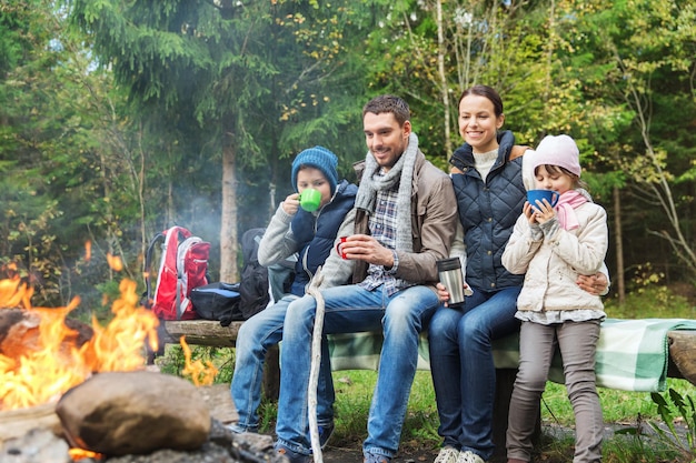 camping, viajes, turismo, caminatas y concepto de personas - familia feliz sentada en un banco y bebiendo té caliente de tazas en el fuego del campamento en el bosque