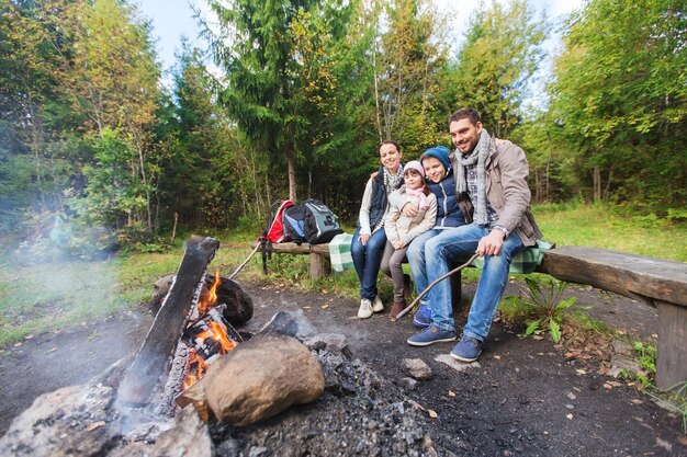 camping, viajes, turismo, caminatas y concepto de personas - familia feliz sentada en un banco en el fuego del campamento en el bosque