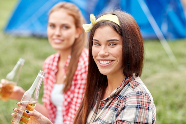 camping, viajes, turismo, caminatas y concepto de personas - mujeres jóvenes felices con botellas de vidrio bebiendo sidra o cerveza en el campamento