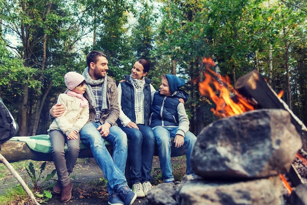 camping, viajes, turismo, senderismo y concepto de personas - familia feliz sentada en un banco y hablando en el campamento cerca de la fogata en el bosque