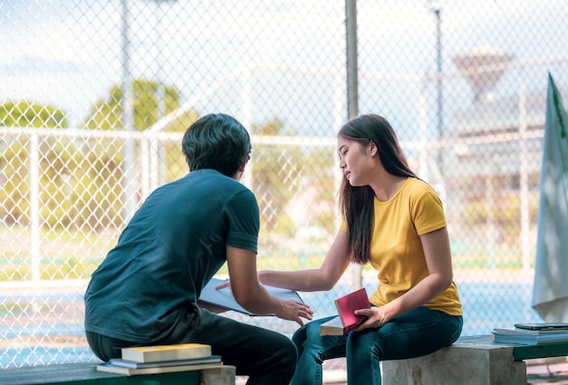 En un campus, un par de estudiantes estudian juntos y un adolescente se sienta en un asiento junto a una cancha de deportes con un libro.