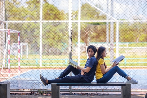 En un campus, un par de estudiantes estudian juntos y un adolescente se sienta en un asiento junto a una cancha de deportes con un libro.