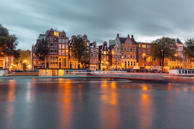 Canal de Amsterdam Amstel con casas típicas holandesas, casa flotante y pista luminosa desde el barco durante la hora azul del crepúsculo, Holanda, Países Bajos.