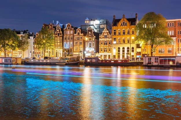 Canal de Amsterdã amstel com casas flutuantes típicas holandesas e trilha luminosa do barco à noite ...