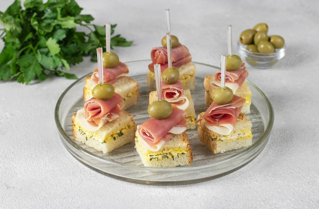 Canapé com jamon e azeitonas em pão branco torrado com queijo sobre fundo cinza claro