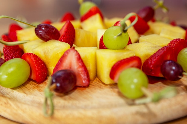 Foto canapés de fruta apetitosa de morango, abacaxi e uvas em uma placa de madeira. fechar-se. buffet de lanches.