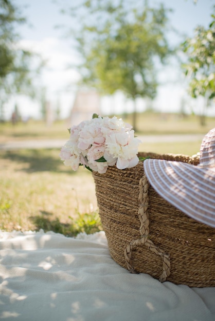 Una canasta con flores, peonías y un sombrero, en el parque.