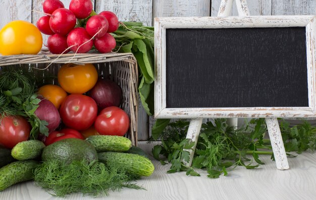 En la canasta hay cebolla, perejil, eneldo, tomates, rábanos, aguacates, pepinos y un pizarrón con copyspace.