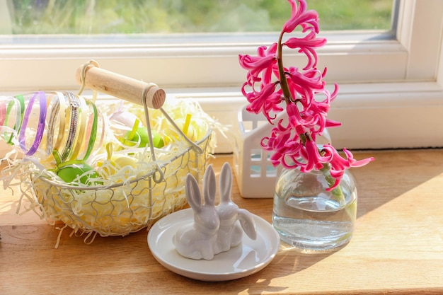 Foto una canasta rústica, conejitos de pascua de cerámica blanca y un jacinto en un jarrón de vidrio sobre un alféizar de madera