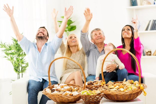 Canastas tejidas con pasteles bellamente decorados, de pie sobre una mesa en la sala de estar. Al fondo se puede ver a una familia feliz sentada en el sofá, con los brazos levantados mirando hacia arriba.