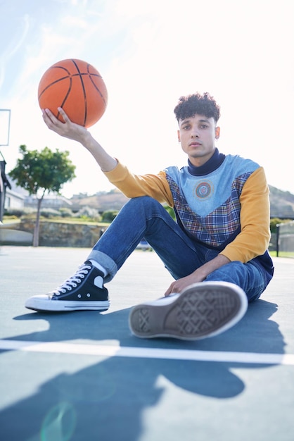 Cancha de baloncesto y retrato de un modelo de hombre sentado en el suelo con ropa elegante y moderna Deportes relajarse y chico de Canadá sosteniendo una pelota en el campo de entrenamiento al aire libre en la ciudad urbana