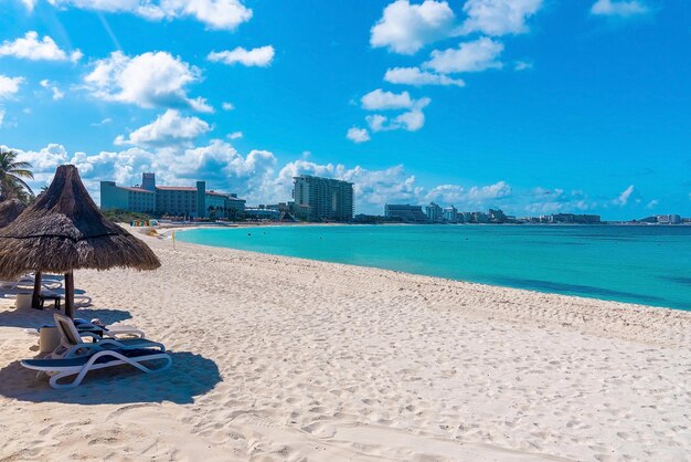 Cancún, méxico. 30 de maio de 2021. espreguiçadeiras vazias sob a cobertura de um telhado de palha na areia da praia com uma linda superfície azul da água do mar e prédios urbanos na costa