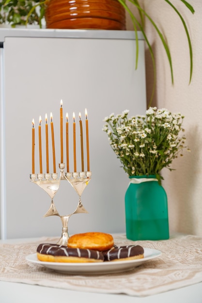 Candelabro de Hanukkah con todas las velas y donas encendidas y un jarrón de flores sobre la mesa. Foto vertical