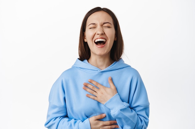 Cándida mujer feliz con sudadera con capucha riendo a carcajadas, sosteniendo las manos en el vientre y el pecho mientras se ríe de algo divertido, broma hilarante, de pie sobre una pared blanca