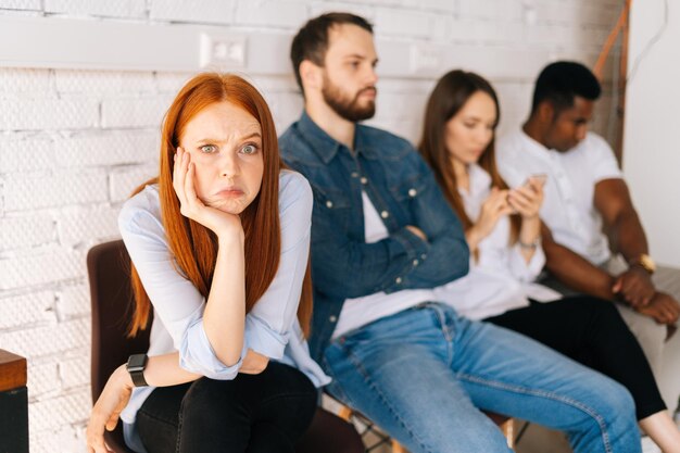 Candidatas cansadas y emocionadas mirando la cámara mientras esperan una entrevista con hr sentadas en fila con candidatas multiétnicas diversas y aburridas