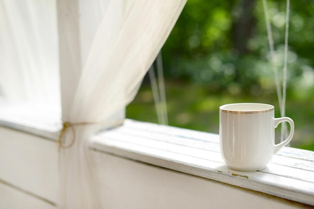 Caneca branca com chá ou café em uma mesa de madeira branca no terraço de verão no fundo do jardim verde