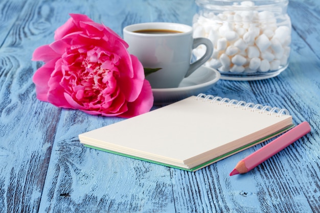 Caneca de café da manhã, caderno vazio, lápis e flores de peônia branca na mesa de madeira azul