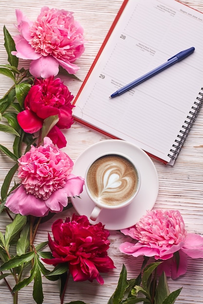 Caneca de café de manhã no café da manhã, caderno vazio, lápis e flores de peônia rosa na vista superior da mesa de pedra branca em estilo liso leigo.