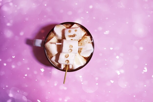 Caneca de cerâmica com bebida quente de cacau fresco e boneco de neve de marshmallow engraçado feito à mão derretido. Efeito de neve. Fundo rosa suave.