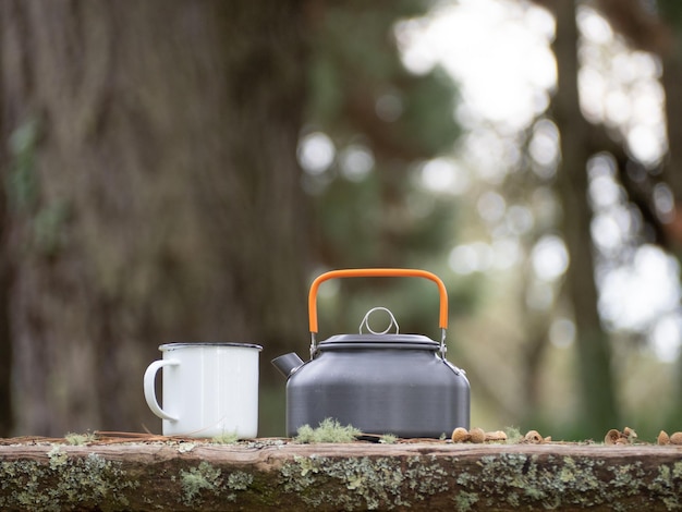 Caneca de esmalte branco com pequeno bule de metal na mesa de madeira Bebida quente lá fora na floresta Caminhada acampando ao ar livre