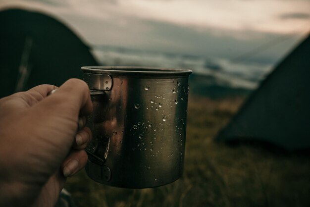 Caneca de metal com chá na mão de um turista em acampamento nas montanhas sob nuvens brancas