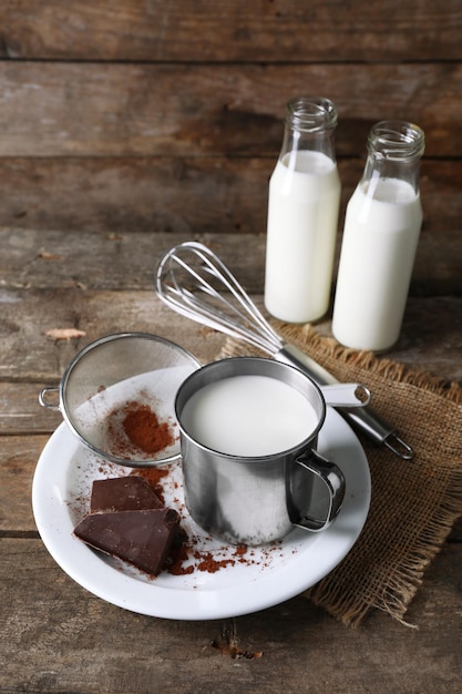 Foto caneca de metal e garrafas de vidro de leite com pedaços de chocolate e coador de cacau no prato com pano de serapilheira e fundo de pranchas de madeira rústica