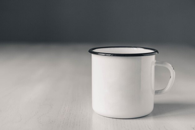 Caneca esmaltada de metal branco com linha preta na borda da mesa de madeira branca sobre fundo cinza