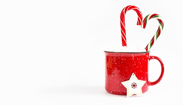 Caneca vermelha com marshmallows e bengala de caramelo em forma de um coração em um fundo branco e uma árvore de natal brinca uma estrela de madeira. natal, ano novo, clima festivo. copie o espaço