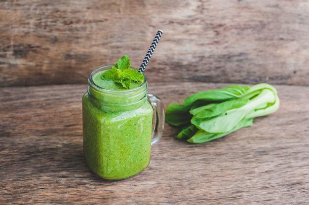 Canecas em frasco de vidro com espinafre verde, banana e batido de leite de coco com uma colher de aveia na mesa de madeira