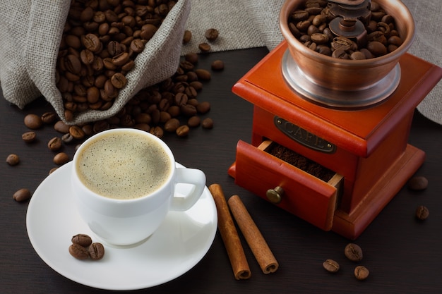 Canela, granos de café y un molinillo de café alrededor de una taza de café.